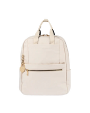 An off-white canvas backpack with gold-tone hardware, featuring dual top handles, a front zippered pocket, and a small heart-shaped charm, shown against a plain white background.