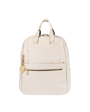 An off-white canvas backpack with gold-tone hardware, featuring dual top handles, a front zippered pocket, and a small heart-shaped charm, shown against a plain white background.